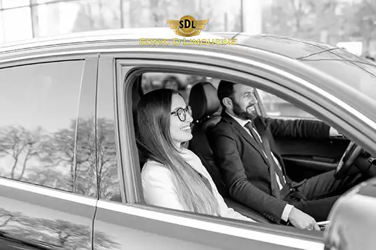 Sonic D Limousine Worldwide Voted Most Reliable Airport Transportation Provider! Black and white image of a smiling woman and a man in business attire sitting in the front seats of a car. The man is driving, and the car has a logo "SDL" with the text "Sonic D Limousine: Service de Luxe. Sonic D Limousine Worldwide your luxury Airport Transportation