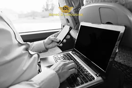 Sonic D Limousine Worldwide Voted Most Reliable Airport Transportation Provider! A person uses a smartphone and laptop while seated on a bus. The "Sonic D Limousine" logo is visible on the window, signifying luxurious airport transportation. The black and white image captures the essence of modern connectivity during travel. Sonic D Limousine Worldwide your luxury Airport Transportation