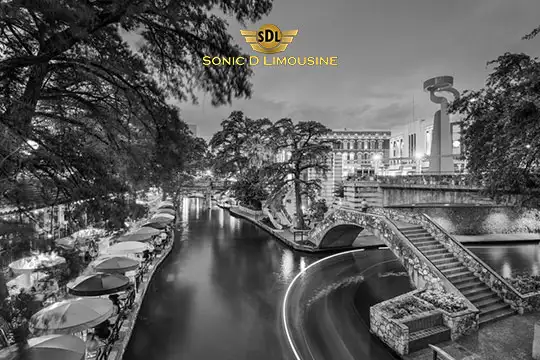 Sonic D Limousine Worldwide Voted Most Reliable Airport Transportation Provider! A nighttime river scene with illuminated bridges and buildings, lined with outdoor tables under umbrellas. Logo of Sonic D Limousine Luxury Airport Transportation at the top center of the image. Sonic D Limousine Worldwide your luxury Airport Transportation