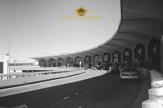 Sonic D Limousine Worldwide Voted Most Reliable Airport Transportation Provider! Black and white photo of vehicles parked near the curved exterior of a large, modern airport terminal. The SDL logo, representing Sonic D Limousine Luxury Airport Transportation, is displayed at the top of the image. Sonic D Limousine Worldwide your luxury Airport Transportation