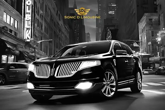 Sonic D Limousine Worldwide Voted Most Reliable Airport Transportation Provider! A black and white image showing a luxury car on a street with urban buildings in the background. The car, part of Sonic D Limousine's fleet, has prominent grille bars and is illuminated by its headlights. The logo "Sonic D Limousine" is above. Sonic D Limousine Worldwide your luxury Airport Transportation