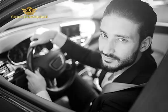 Sonic D Limousine Worldwide Voted Most Reliable Airport Transportation Provider! Black-and-white photo of a man with a beard sitting in a Sonic D Limousine, looking towards the camera, with his left hand on the steering wheel. Sonic D Limousine Worldwide your luxury Airport Transportation