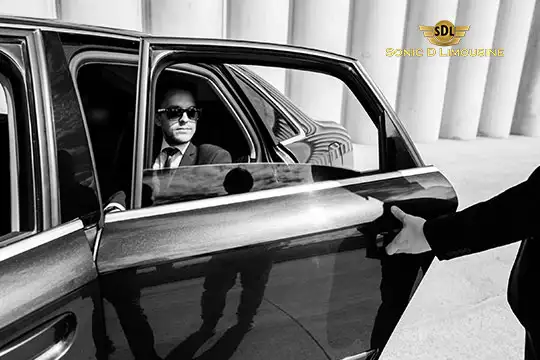 Sonic D Limousine Worldwide Voted Most Reliable Airport Transportation Provider! A person in a suit and sunglasses is seated in the back seat of a Sonic D Limousine with the door open, while another individual holds the door from outside. Sonic D Limousine Worldwide your luxury Airport Transportation